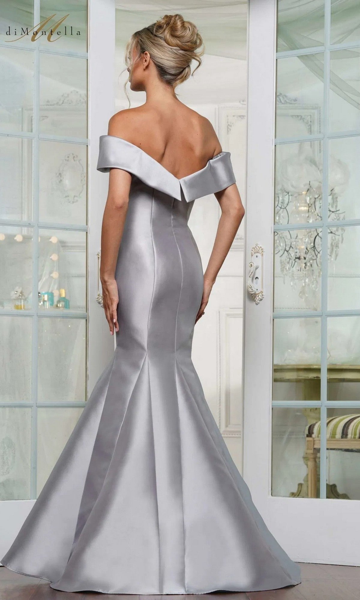 A woman wearing the Long Formal Dress: Rina Di Montella RD3257, a silver off-the-shoulder mermaid gown, stands with her back to the camera in an elegant room with glass doors and a chandelier.